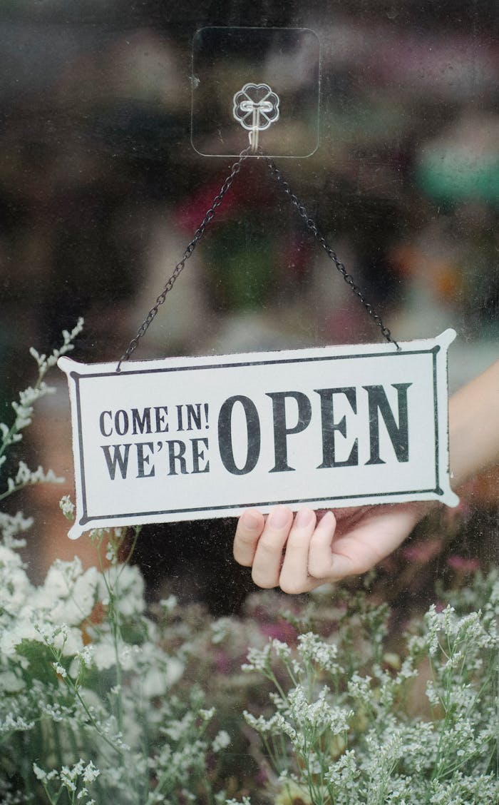 creative-03 Crop anonymous person turning white signboard with inscription Come In We Are Open hanging on glass wall above delicate white flowers